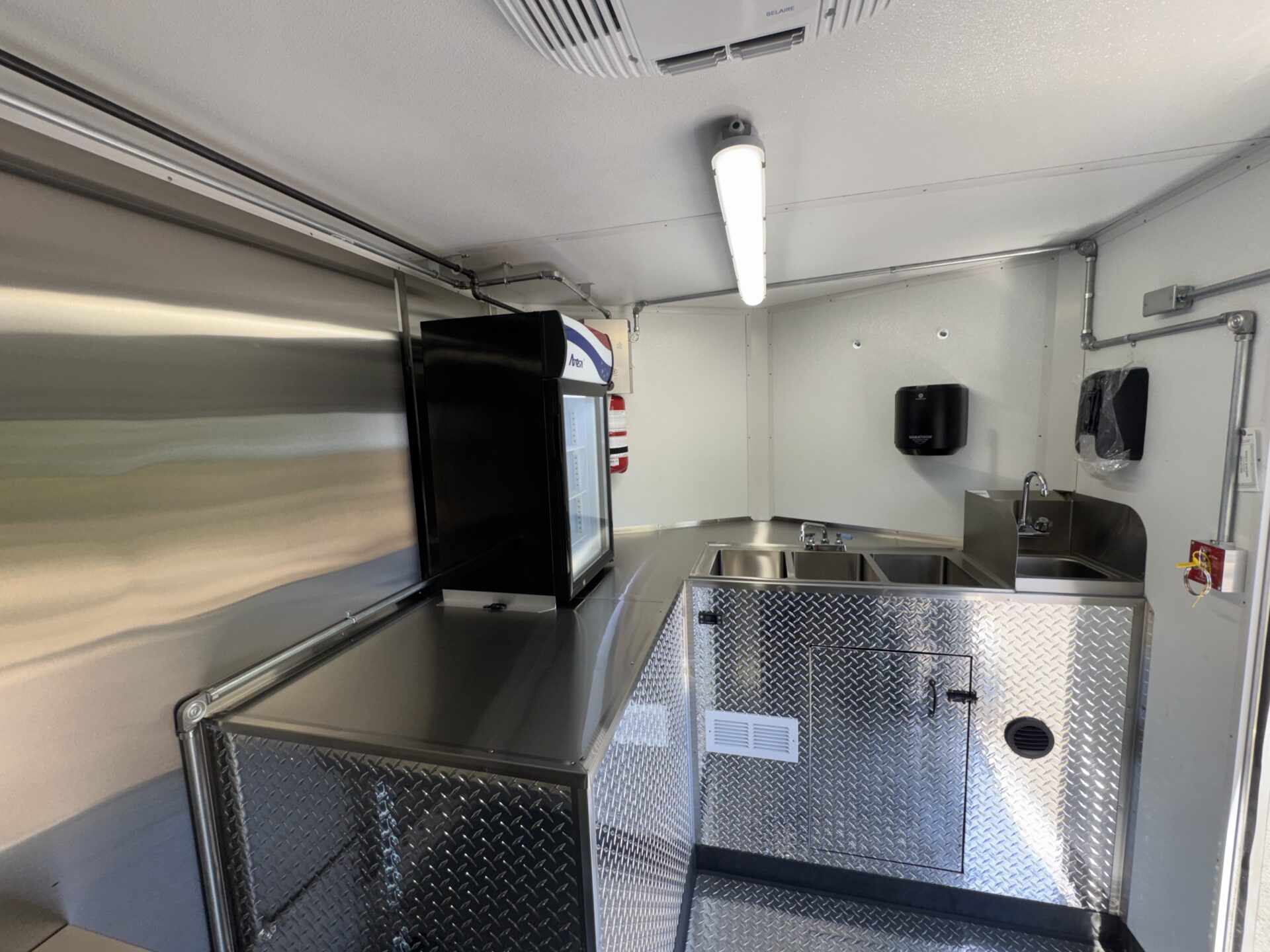 Interior view of the bagel trailer prep area with stainless steel walls, sandwich prep table, merchandiser refrigerator, and three compartment sink