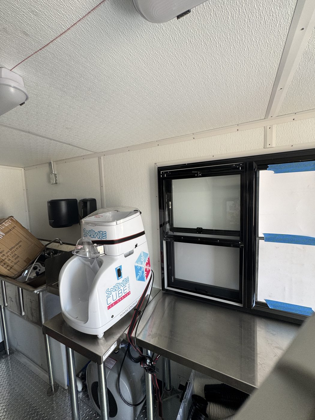 Snow cone machine on a stainless table beside the self closing service window inside the roasted corn food truck